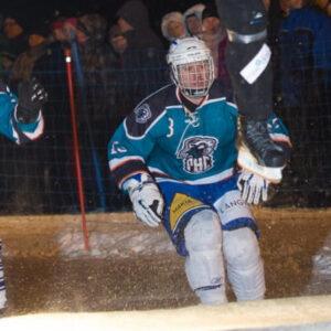 Two ice cross players in blue jerseys watch as a player jumps in front of them on an outdoor rink. A crowd is visible in the background.